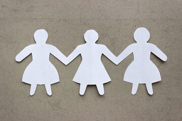 Figures of three girls in dresses holding hands, cut out of white paper. In the center of the photo on a beige background