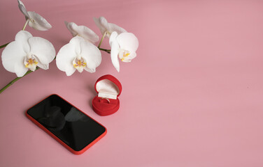 New smartphone and engagement ring in red gift box next to orchid branch on pink background. The concept of best gift for Valentine's Day, birthday, International Women's day and other holidays. 