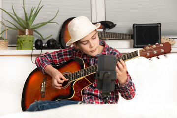 Obraz premium Boy learning or takes off the vlog to play guitar through a video call, home music lessons, hobby, vlogging and creation concept. Young guitarist guy videobloger play the guitar in living room