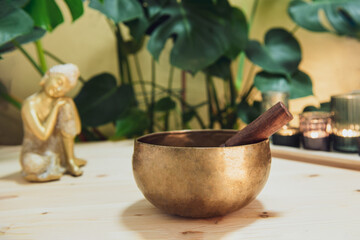Tibetan singing bowl with special stick, burning candles and Golden Buddha decorative statuette on the wooden table with green plants background. Meditation and Relax. Exotic massage. Selective focus