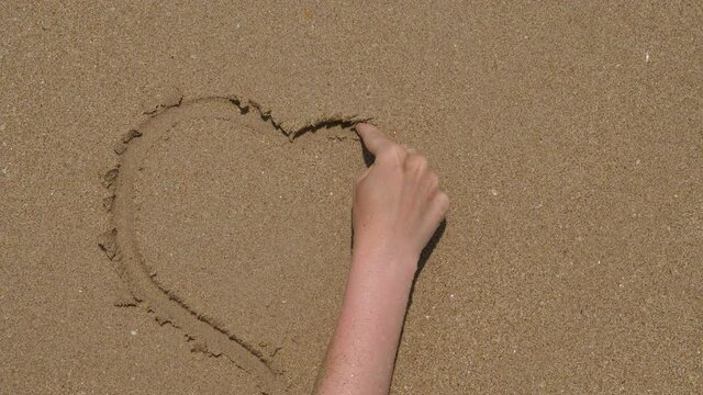 Romantic tourist woman draw heart shape on sand, move finger on wet soft surface. Sea waves surf and rush on beach, wipe fragile image away, top-down close shot