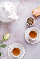 Two porcelain cups of herbal tea,teapot,cakes and beautifull white rose on white marble background.Top view.