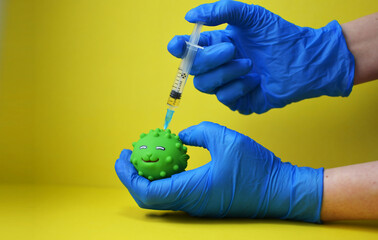 A woman wearing medical gloves gives an injection of the covid 19 coronavirus vaccine to a toy. A toy virus and a syringe on a yellow background. The medical concept of combating the covid-19 pandemic