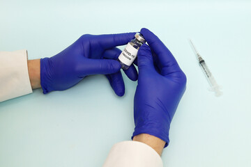 A bottle of Covid-19 vaccine in the hands of a doctor wearing blue medical gloves and a syringe on a blue background. Concept: vaccination against coronavirus