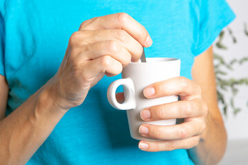 hands of middle-aged woman in blue t-shirt stirring the teaspoon in a white cup of coffee or tea