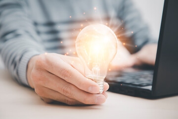 businessman hand holding light bulb and working with computer on the desk, saving energy and...
