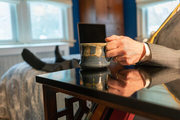 Working from home man with laptop sits on bed holding cup of coffee wearing suit, tie and with stocking feet, humor