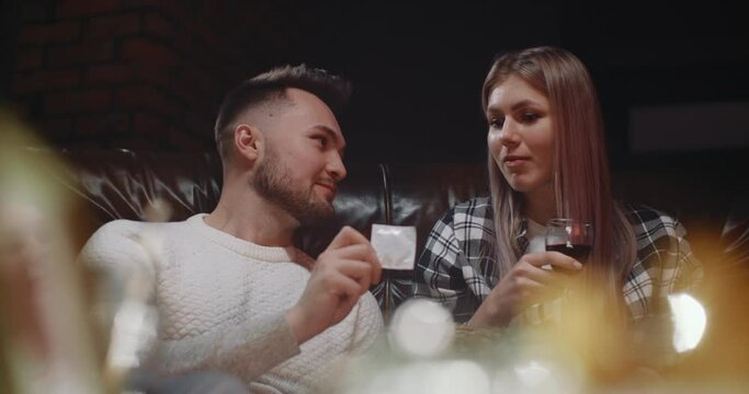 Millennial couple in love having fun watching TV eating snacks drinking wine at home. Young man taking condom and show it playfully offers sex to his girlfriend. Concept of sexual relationships