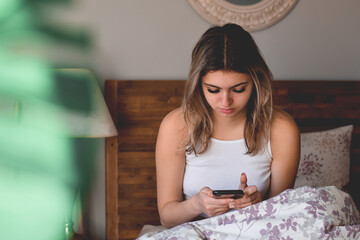 Young teenage girl sitting in bed just woken up using mobile phone texting