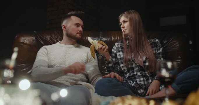 Millennial couple having fun watching TV. Young female eating banana, her boyfriend looks at her, flirts, takes condom and show it playfully offers sex to girlfriend. Concept of sexual relationships