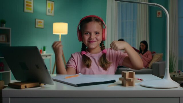 Energetic schoolgirl in pink headsets is sitting at desk and has fun time during online class, dancing and smiling at camera, enjoying distant study, while parents relax on sofa, Zoom out, Slow motion