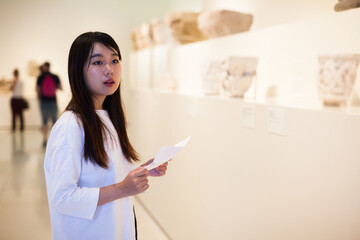 Portrait of chinese girl holding guidebook, standing in museum of ancient sculpture