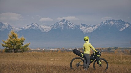 Fototapeta premium The woman travel on mixed terrain cycle touring with bikepacking. The traveler journey with bicycle bags. Sport tourism bikepacking, bike, sportswear in green black colors. Mountain snow capped.