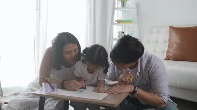 Happy Asian family at home. Parents with little daughter sit on the floor using color pencil drawing on paper book. Father and mother with cute child girl kid having fun leisure homeschooling together