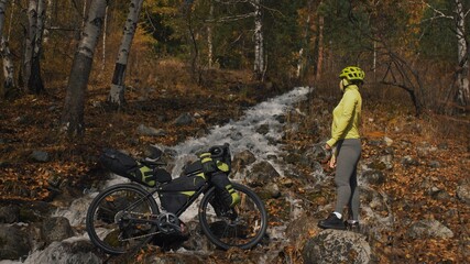 Obraz premium The woman travel on mixed terrain cycle touring with bikepacking. The traveler journey with bicycle bags. Sportswear in green black colors. The trip in magical autumn forest, river stream.