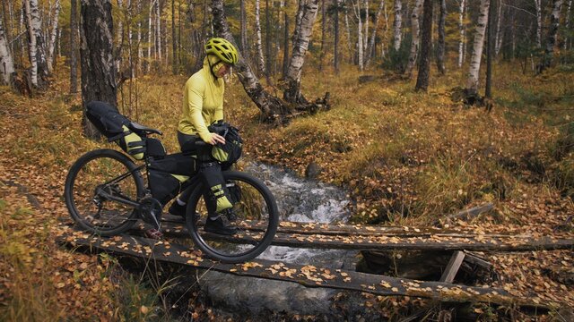 The woman travel on mixed terrain cycle touring with bikepacking. The traveler journey with bicycle bags. Sportswear in green black colors. The trip in magical autumn forest. Wooden bridge, river.