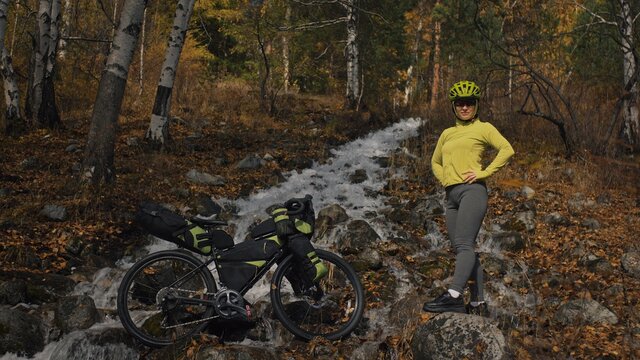 The woman travel on mixed terrain cycle touring with bikepacking. The traveler journey with bicycle bags. Sportswear in green black colors. The trip in magical autumn forest, river stream.