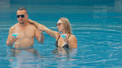 Obraz na płótnie Canvas The loving couple hugs and kisses, drinking blue cocktail alcohol liquor in swimming pool at hotel outdoor. Portrait of caucasian man and woman. Creative hairstyles bodybuilder, swimsuit, sunglasses.
