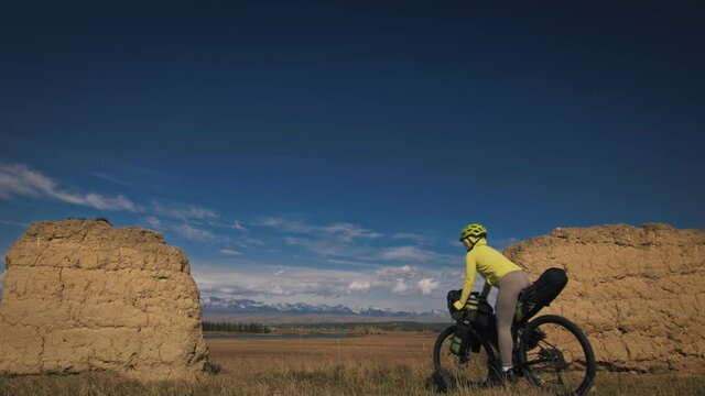 The woman travel on mixed terrain cycle touring with bikepacking. The traveler journey with bicycle bags. Sport bikepacking, bike, sportswear in green black colors. Mountain snow capped, stone arch.