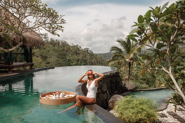 Young pretty woman in white swimsuit siy on the edge of infinity pool surrounded wth tropical nature and coconut palm trees, enjoy floating breakfast and idyllic morning on Bali island