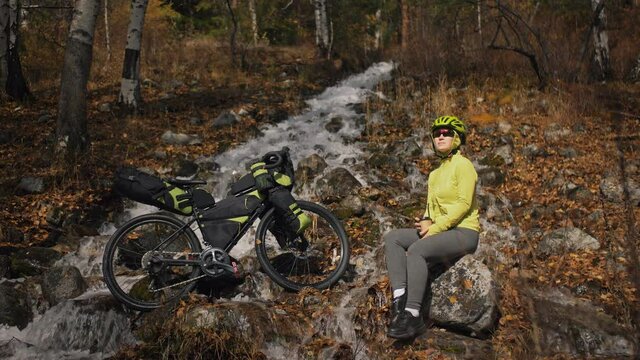 The woman travel on mixed terrain cycle touring with bikepacking. The traveler journey with bicycle bags. Sportswear in green black colors. The trip in magical autumn forest, river stream.