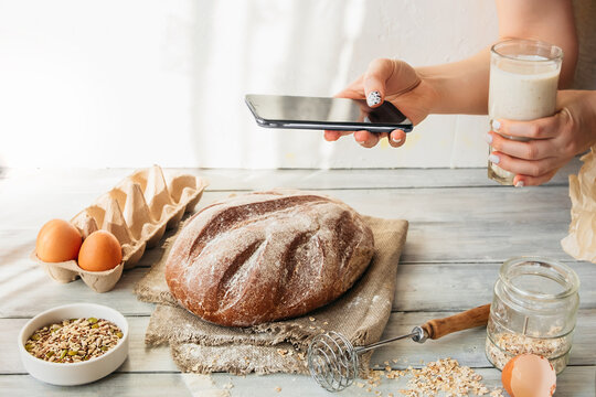 unrecognizable person takes pictures of cooking homemade food on smart phone. homemade baked grain bread with seeds. kitchen table with eggs, flour and pastries. food blogging, online cooking at home