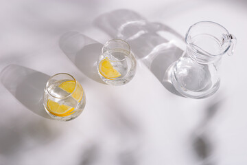 Drinking natural water in beautiful glass glasses with slice of lemon inside on white background in sunny day. Summer minimal flat lay with hard shadows.