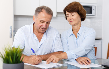 Fototapeta premium Portrait of positive mature couple at kitchen table filling up documents