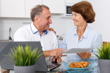 Portrait of smiling mature family couple with documents using laptop at kitchen table