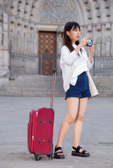 Young chinese woman traveler strolling with luggage around city, making photo of sights. High quality photo