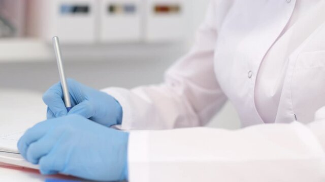 4k Unrecognizable woman doctor surgeon in white uniform is writing diagnosis and examination results. Hands in blue gloves close up. Healthcare and medicine
