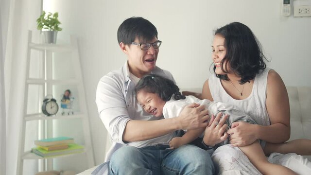 Happy Asian family relaxing at home. Smiling parents embracing and playing with little daughter on sofa in living room. Father and mother with cute child girl kid having fun weekend activity together.