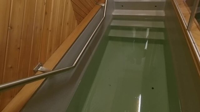 closeup steps with metal railing leading into a baptismal pool with wooden siding inside a church with a plexiglass seperation and the water reflecting people entering to participate in ceremony 2-2