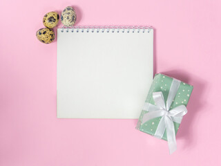 Blank sheet of paper for text surrounded by quail eggs and gift box on pink background. Preparation for easter. Happy Easter Holiday card with space. Easter shopping list. Easter holiday concept.