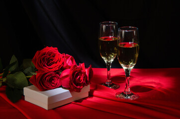 Glasses with champagne, red roses and a gift on a red tablecloth