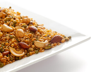 'Shahi Mixture' in a white ceramic square plate, made with peanut, cashew, corn flakes, almonds, etc. Pile of Indian spicy snacks (Namkeen). Side view, against the white background