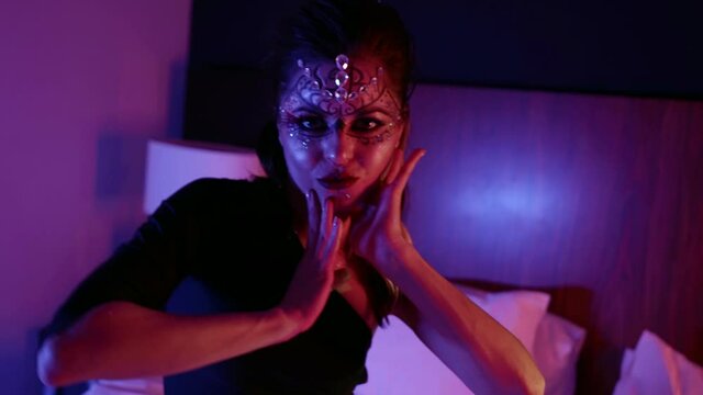 A woman with unusual makeup, a pattern and with rhinestones on her face poses in front of the camera in the studio while on the bed. Makes a motion with his hands.