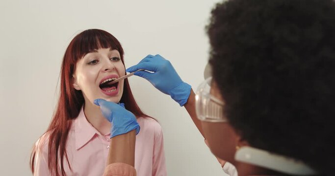 Concept Of Treatment And Prevention Of Throat Disease. An Otolaryngologist Afro American Doctor Woman Examines A Female Throat With A Wooden Spatula. A Possible Diagnosis Is Inflammation