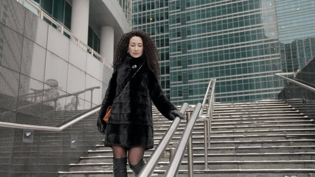 A curly-haired, stylish, middle-aged brunette woman in a black fur coat walks alone in the city on a cold day. He smiles and goes down the stairs near the building.