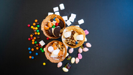 Ice cream in waffle cups, decorated with sweets, marshmallows and nuts on a dark background. Background surface with copy space for text.