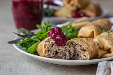 Homemade stuffed thin pancakes or crepes with meat, spicy cranberry sauce and arugula, light stone background. Traditional Russian cuisine for Maslenitsa. Shrove Tuesday.