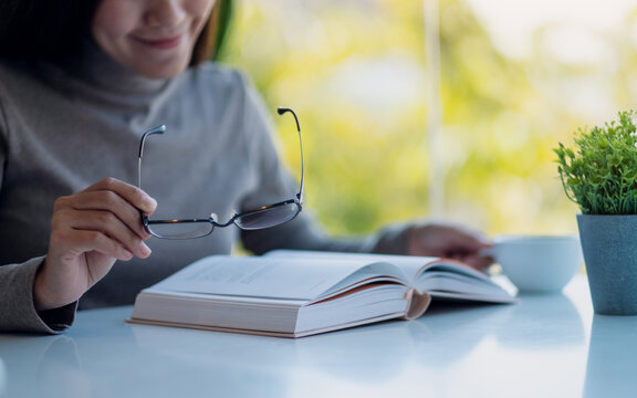 Closeup image of a beautiful young asian woman holding eye glasses while reading book