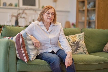 Portrait of an elderly woman sitting on a couch at old age home. Caucasian woman sitting on a sofa and looking at camera. Care for lonely pensioners.