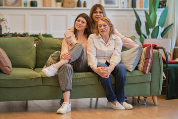 three generations of a family of women are sitting together on the couch. grandmother, daughter, and granddaughter pose together to get a joint photo.