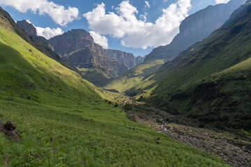 Fototapeta na wymiar Hikers in lush valley below the towering escarment of the Drakensberg Mountains.