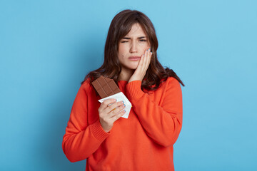 Woman suffering from toothache after eating chocolate, frowning her face, covering cheek with palm, female with dark hair wearing orange casual sweater.