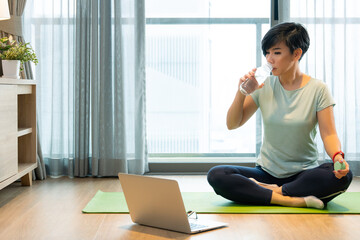 Home exercise during Covid 19 lock down. A healthy asian woman sit cross legs on a yoga mat, sip...