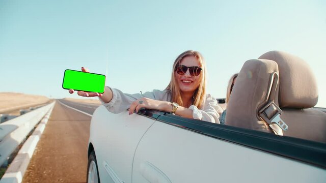 A woman with glasses on a convertible shows her phone and smiles. The scene takes place on the highway. The weather is clear. A woman sits with friends in a car and holds a phone with a green screen