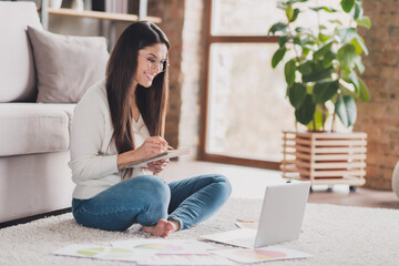Photo of pretty young girl sit floor write notepad look netbook wear eyewear white pullover in living room home indoors
