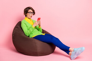 Profile side photo of excited woman wear green cardigan spectacles sitting bean bag chatting device isolated pink color background